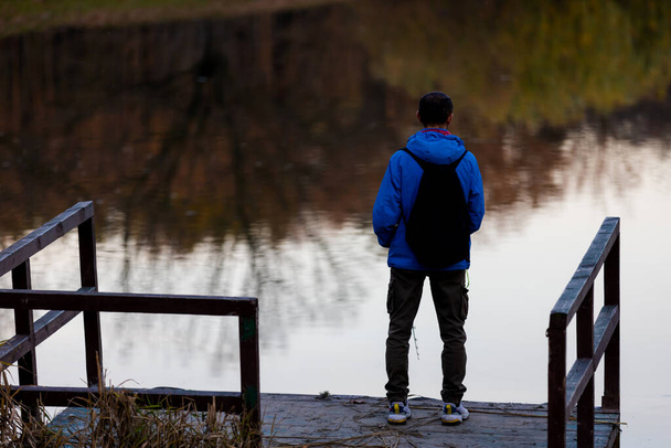 Mladý muž s batohem, který stál sám na dřevěné lávce a zíral na jezero. Klidná atmosféra v přírodě. Užívá si čerstvého vzduchu na podzim večer. Zadní pohled. - Fotografie, Obrázek