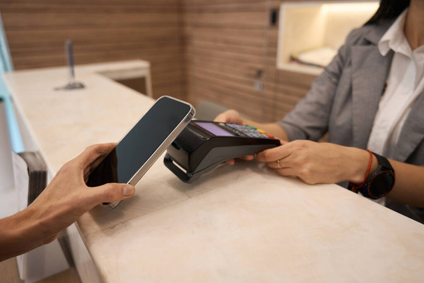 Woman hand holding mobile phone paying the bill with contactless technology at the reception desk - Foto, Imagem