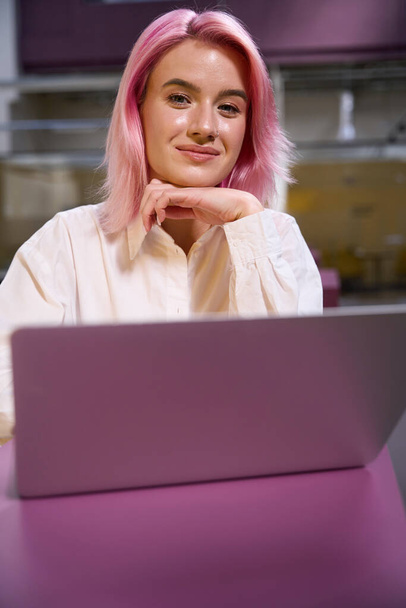 Όμορφη γυναίκα με ροζ μαλλιά κάθεται δίπλα στο τραπέζι με φορητό υπολογιστή σε συνεργασία και κοιτάζοντας την κάμερα - Φωτογραφία, εικόνα