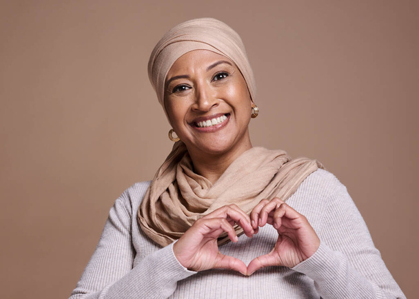 Handen, hart en moslim met een volwassen vrouw in de studio op een bruine achtergrond om liefde of gezondheid te bevorderen. Portret, handteken en islam met een oudere vrouw die poseert voor gezondheidszorg, affectie of cultuur. - Foto, afbeelding