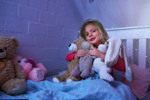 Yatma zamanı küçük dostlarım. Küçük bir kızın portresi. Yatakta otururken yumuşak oyuncaklarını kucaklıyor. - Fotoğraf, Görsel