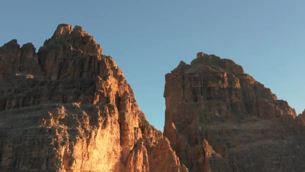 Scherpe gezichten van Drie pieken van Lavaredo onder heldere blauwe hemel verlicht door de rijzende zon. Landschap van majestueuze plantenloze bergtoppen bij zonsopgang vanuit de lucht - Video