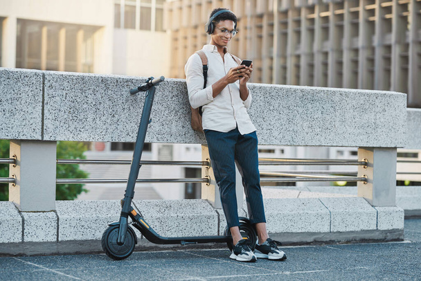 Telefoon, muziek en elektrische scooter met een zakelijke zwarte man die audio streamt tijdens zijn woon-werkverkeer. Mobiel, koptelefoon en vervoer met een mannelijke werknemer die een sms-bericht typt in een stedelijke stad. - Foto, afbeelding