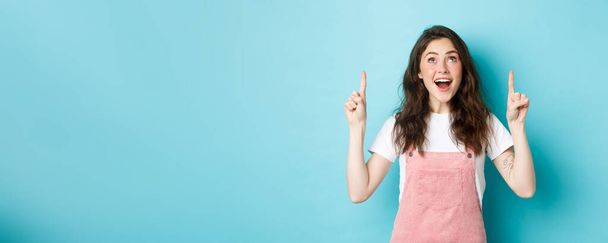 Portrait of excited brunette girl in summer outfit, looking and pointing fingers up with happy face, checking out promo offer, showing cool advertisement, blue background. - Фото, изображение