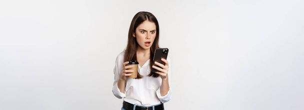 Woman holding coffee cup and looking at smartphone with shocked, stunned face, standing over white background. Copy space - Foto, imagen