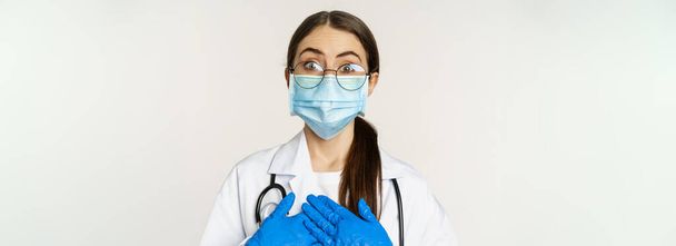 Portrait of surprised and amazed young doctor, looking with fascination, holding hands on heart, standing in medical face mask over white background. - Fotoğraf, Görsel