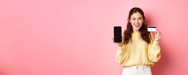Τεχνολογία και online αγορές. Νεαρό ελκυστικό θηλυκό μοντέλο δείχνει άδειο οθόνη smartphone με πλαστική πιστωτική κάρτα, δείχνουν τραπεζικό λογαριασμό ή εφαρμογή, στέκεται πάνω από ροζ φόντο. - Φωτογραφία, εικόνα