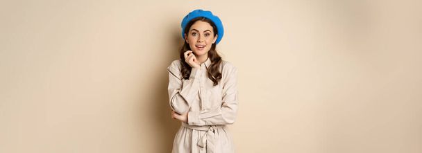Image of stylsh beautiful woman looking surprised, shocked reaction at camera, posing in trench coat against beige background. - Fotografie, Obrázek