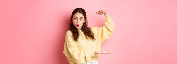 Surprised young woman holding big empty space between her hands, showing large size and staring impressed at camera, standing against pink background. - Photo, image
