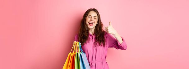 Молода щаслива жінка-покупець, яка показує великі пальці вгору, задоволена хорошими знижками, купує персонал на продаж, тримає сумки для покупок і посміхається задоволений, рожевий фон
. - Фото, зображення
