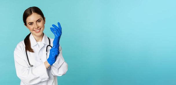 Smiling young woman, medical worker wearing gloves for patient checkup, standing in hospital clinic uniform over blue background. - Photo, Image