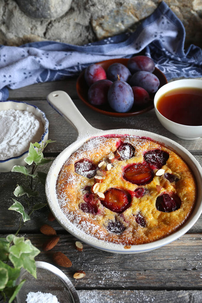 Damson blue plum clafoutis (flan) and cup of tea, icing sugar and almonds dressing, french cuisine on wooden background. Rustic style - 写真・画像