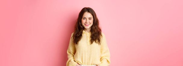Retrato de boa aparência jovem mulher em suéter, de mãos dadas em bolsos, sorrindo e olhando determinado para a câmera, de pé contra fundo rosa. - Foto, Imagem