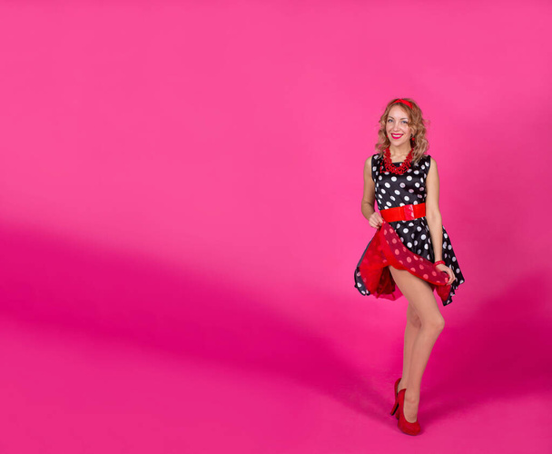 赤いアクセサリーを持つ白いポルカドットと黒のドレスでピンクの背景にブロンドの女の子:ハイヒールの靴、ブレスレットとイヤリングのビーズ、彼女の頭の上にフープ。赤い口紅の唇。ピンナップ. - 写真・画像