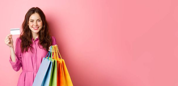 Joven morena sosteniendo bolsas de compras, mostrando tarjeta de crédito de plástico y sonriendo, de pie sobre fondo rosa. - Foto, imagen