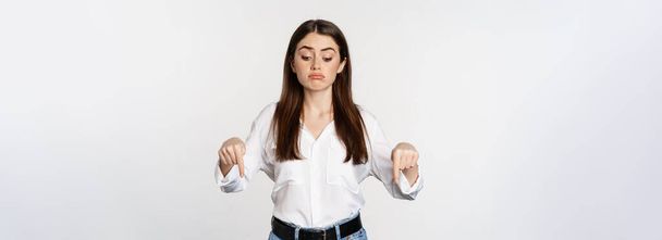 Разочарованная молодая женщина, указывающая пальцами вниз, с сожалением и грустью глядя снизу, стоящая в блузке и джинсах на белом фоне. - Фото, изображение