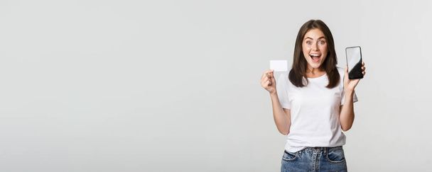 Ενθουσιασμένος και έκπληκτος χαριτωμένο κορίτσι δείχνει πιστωτική κάρτα και κινητό τηλέφωνο τραπεζική εφαρμογή στην οθόνη. - Φωτογραφία, εικόνα