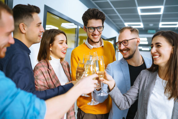 成功のコンセプトに乾杯オフィスに立っている間、若いビジネスの人々がお互いをトーストし、笑顔のグループ。お祝いとお祝いのコンセプト. - 写真・画像