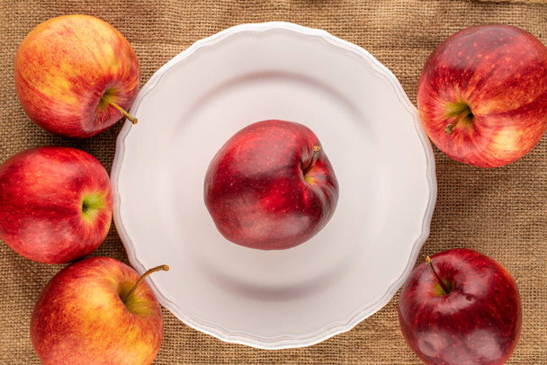 Varias manzanas rojas jugosas con un plato blanco en un paño de yute, macro, vista superior. - Foto, imagen