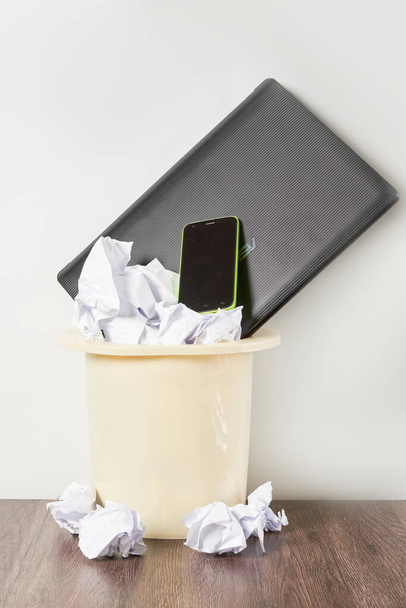 ένα κινητό τηλέφωνο σε ένα σκουπιδοτενεκέ με τσαλακωμένο χαρτί στο πάτωμα δίπλα του είναι ένας ανοικτός υπολογιστής laptop - Φωτογραφία, εικόνα