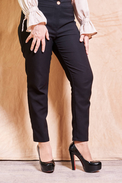 una mujer con las manos en las caderas con pantalones negros y zapatos de tacón alto, de pie frente a un telón de fondo beige - Foto, imagen