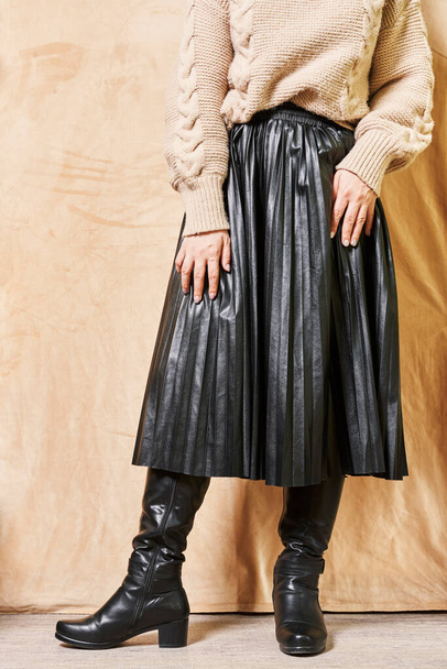 eine Frau in schwarzem Lederrock und kniehohen Stiefeln, die vor einer beigen Wand steht, die Hände auf den Hüften - Foto, Bild