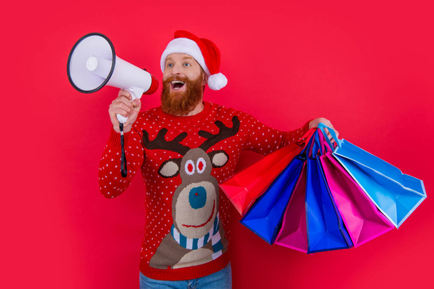Bearded man shouting to loudspeaker holding shopping bags. Santa Claus making discount announcement. Christmas sale announcement. Shop announcement. Merry Christmas. - Photo, image