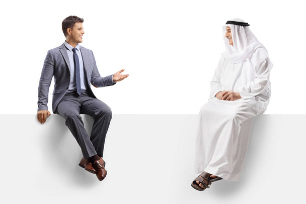 Arab man in an ethnic robe sitting on a blank panel and listening to a businessman talking isolated on white background - Photo, image