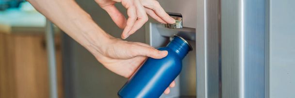 Male hand is pouring cold water and ice cubes in a metal bottle from dispenser of home fridge. BANNER, LONG FORMAT - Фото, изображение