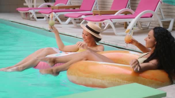 Gyönyörű napozás két lány barátok multietnikus nők érezzék jól magukat a nyári medence frissítő lé jég koktélok ünnepelni nyaralás hotel spa nevetés fröccsenő víz úszás felfújható gyűrűk - Felvétel, videó