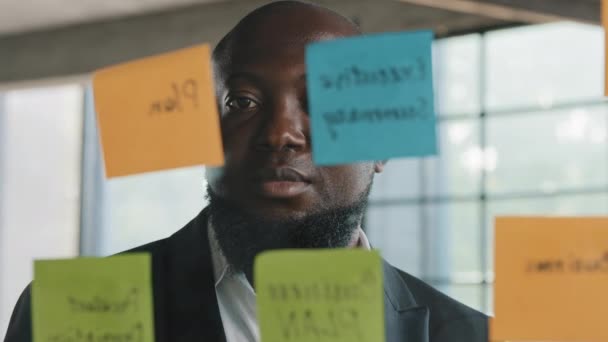 Afrikai amerikai vezető középkorú szakember üzletember vállalkozó főnök munkáltató tervező állni a modern irodában üveges fal etnikai férfi munkás olvasni matricák jegyzetek szervezni menedzsment folyamat - Felvétel, videó