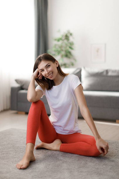 Stunning young woman in sport clothes sitting on floor and smiling sincerely. Caucasian dark haired lady feeling satisfaction after morning workout at home. - Фото, изображение