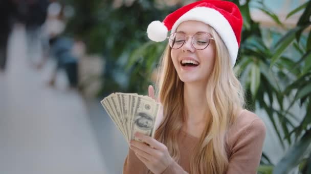 Щаслива багата кавказька жінка в червоному капелюсі Санта-Різдва і окуляри тримати гроші в руках рахуючи грошовий приз успіх перед покупками фінансова знижка до Нового року показують пальцем вгору - Кадри, відео