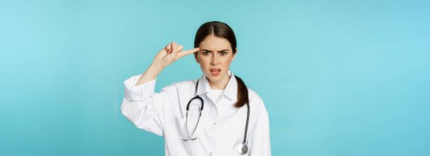Médico mujer molesta señalando con el dedo a la cabeza, regañando a alguien estúpido, loco o extraño, de pie sobre fondo de color turquesa. - Foto, imagen