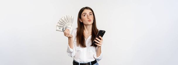 Portrait of happy smiling woman using mobile phone app, holding money cash, standing over white background. Copy space - Foto, Imagem