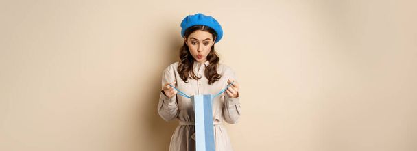 Image of girl looking surprised after opening gift bag, looking at present amazed, standing in stylish outfit over beige background. - Foto, Bild