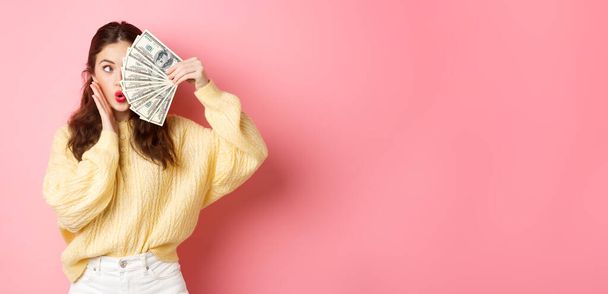 Coquettish nuori nainen pucker huulet, joten typerä kasvot, kattaa puolet siitä rahaa dollareita, seisoo yli vaaleanpunainen tausta. Kopioi tila - Valokuva, kuva
