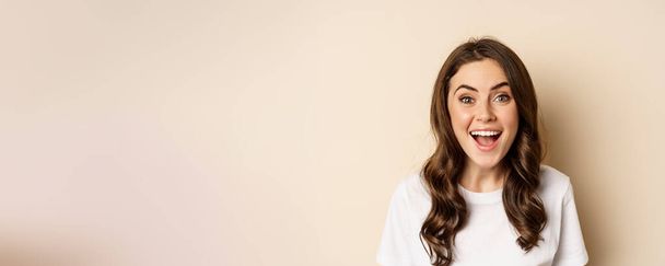 Close up portrait of surprised and excited brunette girl looking amazed, reacting impressed and excited, standing against beige background. - Foto, imagen