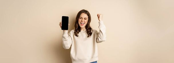 Happy woman winning, showing smartphone screen and shouting of joy, fist pump, standing over beige background. - Foto, Imagem