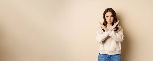 Woman showing stop, cross sign gesture and frowning angry, dislike and disapprove, saying no, rejecting and prohibiting, standing over beige background. - 写真・画像