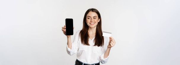 Portrait of female model showing credit card with smartphone screen, recommending application, standing over white background. - Foto, afbeelding