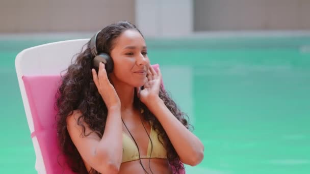 Boldog latin-amerikai spanyol arab nő lány pihenés a strandon fülhallgató hallgatni kedvenc dal zene élvezni nyári nyaralás nyaralás ülni nyugágyon medencével mosolygós Y-generációs hölgy hűtés csukott szemmel - Felvétel, videó