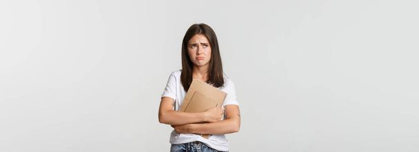 Portrait of sad and disappointed cute female student holding notebooks and looking upper left corner upset. - Photo, Image