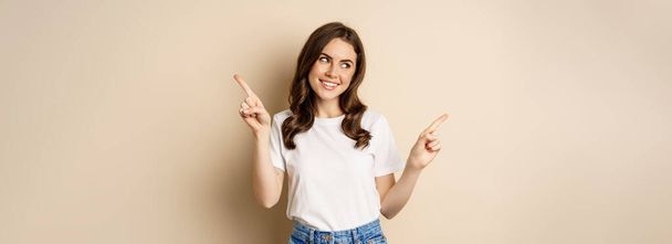 Happy authentic girl smiling, pointing fingers sideways, showing left and right banner, demonstrating promo, standing against beige background. - Foto, afbeelding
