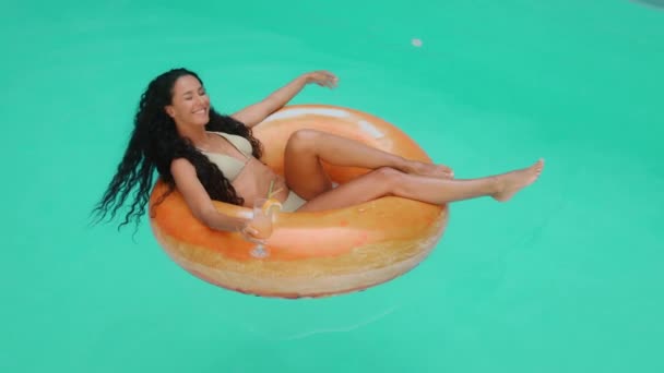 Привлекательная загорелая кудрявая девушка загорает в купальнике в бикини отдыхает в бассейне отеля молодая сексуальная латиноамериканка плавает на надувном спасательном буе с коктейлем чувствовать свободу насладиться летним жарким отпуском - Кадры, видео