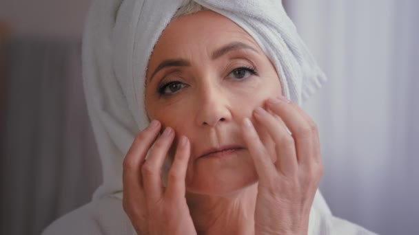 Close up old senior mature Caucasian 50s woman with bath towel on head massaging facial wrinkle touching massage wrinkled face skin care with anti age lifting moisturizing cream healthy complexion - Footage, Video