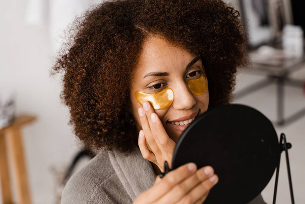 Morning beauty routine. African american woman with golden patches on her eyes looks in the mirror at her skin. African girl in bathrobe applies gold patches to her eyes in the bathroom - Photo, Image
