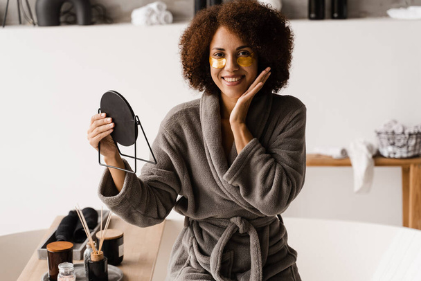 African girl in bathrobe with golden eye patches is looking at the mirror in the bathroom. Morning beauty routine of african american woman - Photo, Image