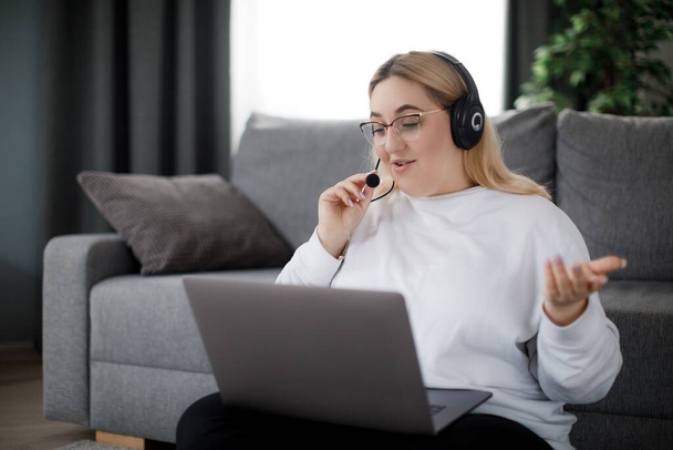 Beautiful young woman with extra weight sitting on floor and using laptop with headset for online education. Concept of modern gadgets and distance learning. - Photo, Image
