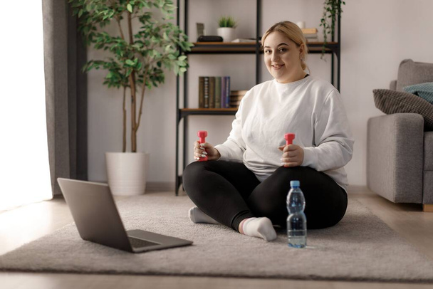 Charming woman in activewear with dumbbells in hands sitting on floor and using modern laptop for online exercises. Caucasian blonde having regular workout at home for losing weight. - Фото, изображение